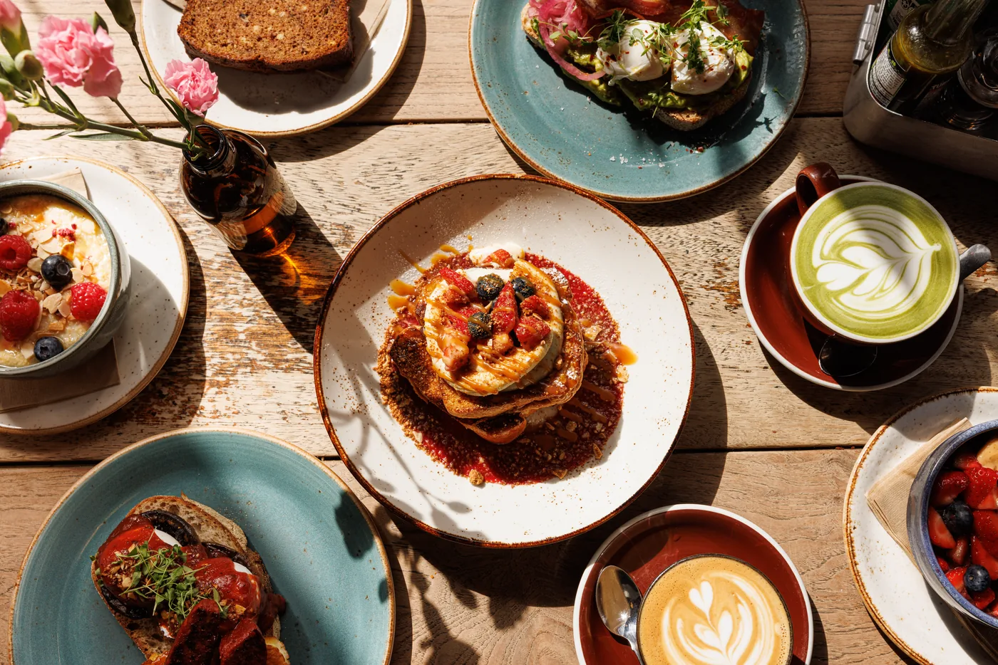 A wooden table shot from above, set with French toast topped with berries, avocado toast with poached eggs, a bowl of porridge with raspberries and blueberries, banana bread, a matcha latte and a cappuccino.