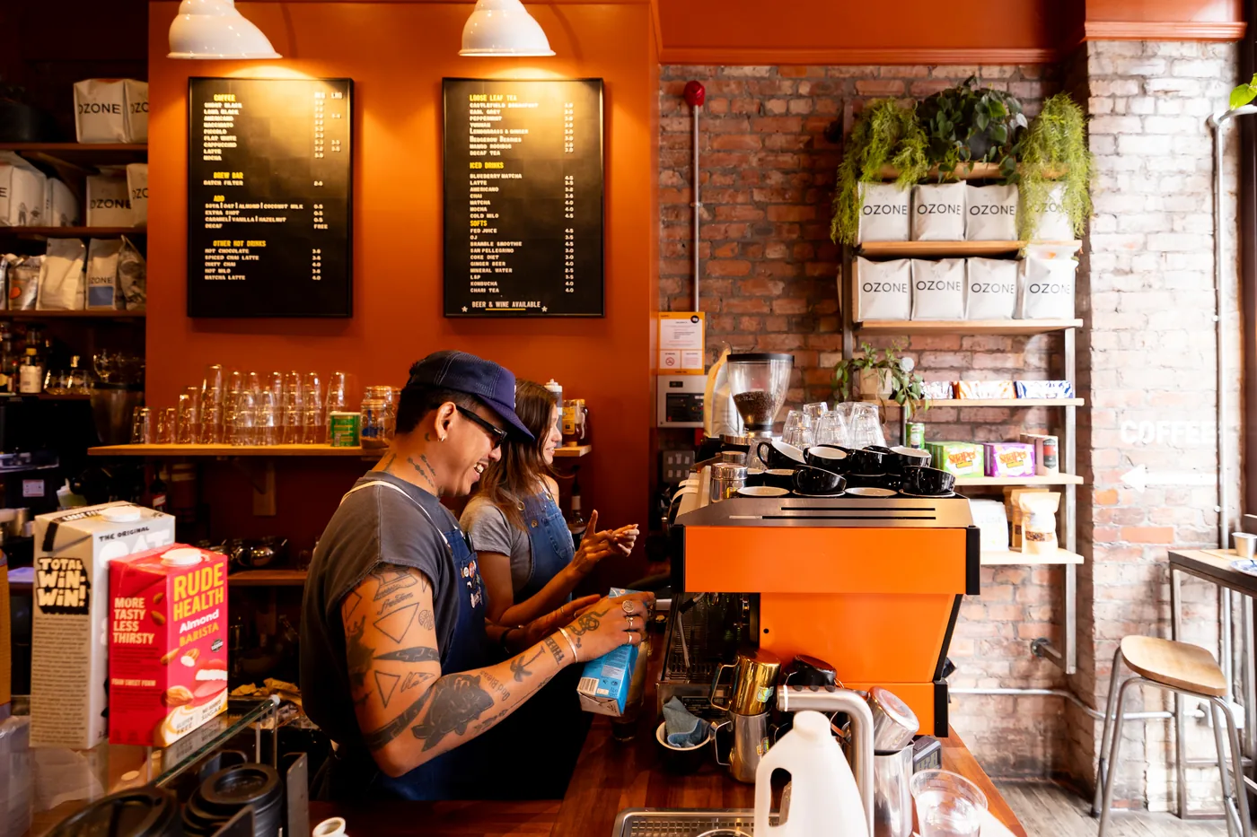 Two members of staff in aprons working behind a coffee bar, next to an orange espresso machine, in a cafe with burnt orange walls, exposed brickwork and chalkboard menus.