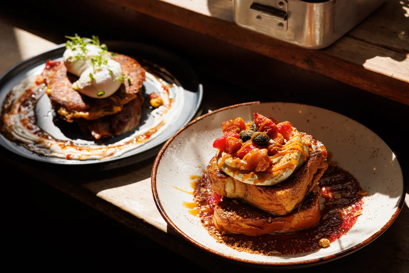 Two plates in low window light: French toast layered with cream, berries and caramel sauce, and behind it a savoury stack topped with a poached egg and micro herbs.