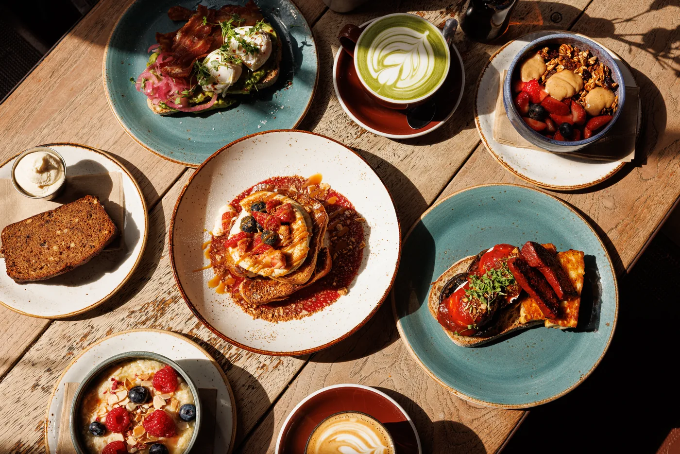 A sunlit wooden table shot from above with French toast and berries in the centre, avocado toast with bacon and poached eggs, a matcha latte, a bowl of berries and granola, banana bread and a bowl of porridge.