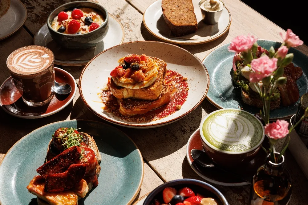 A rustic table laid with French toast topped with berries, a hot chocolate with latte art, a matcha latte, avocado toast with bacon, a bowl of fruit and a small vase of pink carnations.