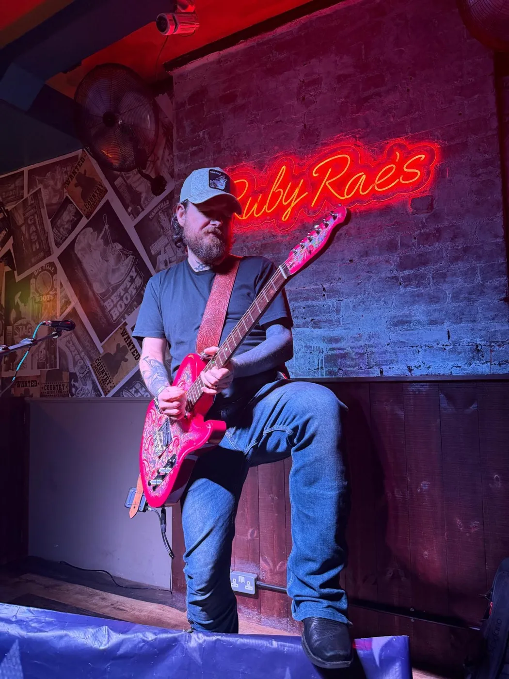 A guitarist in a cap playing a red guitar on a small stage under a pink neon sign reading Ruby Rae's